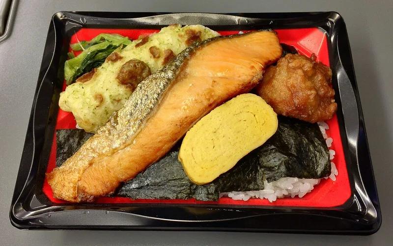 A bento box with grilled salmon, rolled omelette, fried chicken and rice.